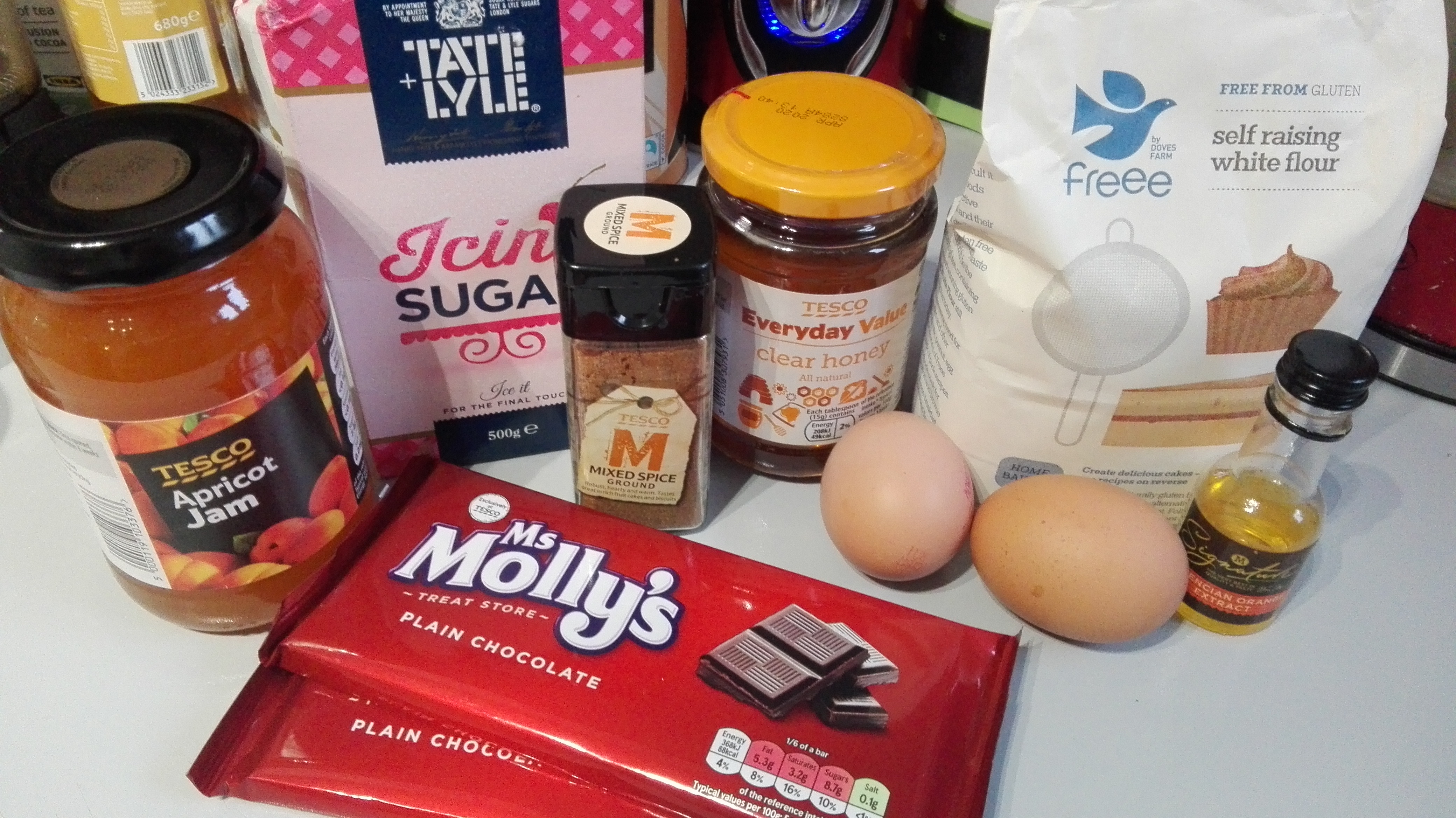 gluten free lebkuchen ingredients - apricot jam, icing sugar, mixed spice, honey, 2 eggs, gluten free flour and 2 bars of dark chocolate on a kitchen worktop