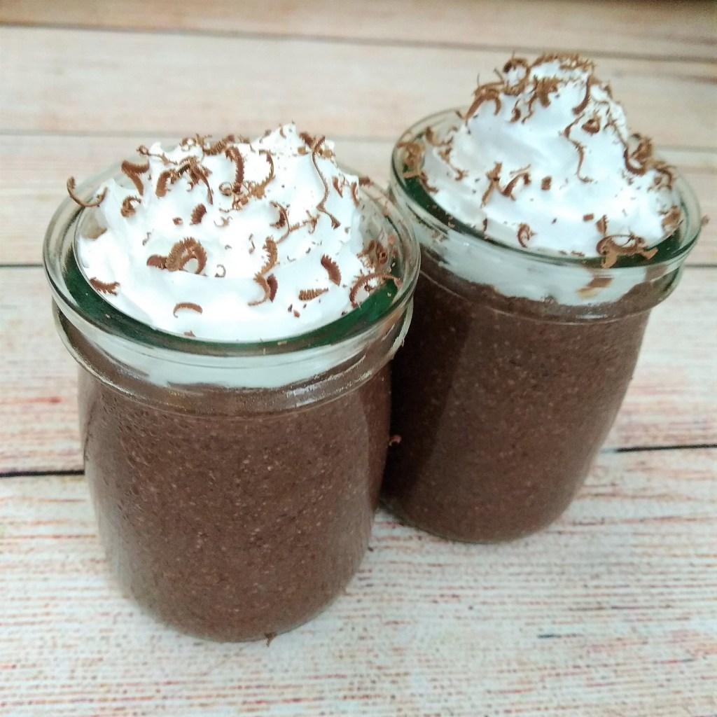 smooth chocolate cherry chia pudding topped with vegan squirty cream and chocolate shavings in glass jars on a table