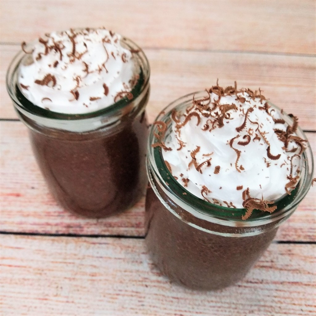 two glass jars of dairy free vegan chocolate cherry pudding topped with whipped cream and chocolate shavings on a table.